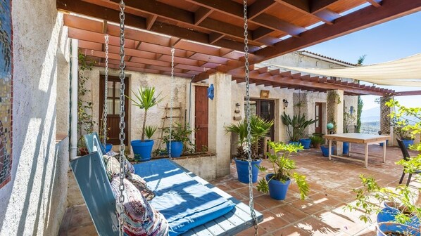 Un espacio exterior cubierto con asientos, una silla colgante, plantas en macetas y una mesa.
