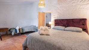 A bedroom with a large bed, a smaller bed, a wooden side table, a hanging light fixture, and a door leading to another room.