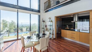 A modern dining area with a glass table and chairs, a kitchen with wooden cabinets, and a large window offering a view of a swimming pool and greenery. - Idem Rural Baza by Ruralidays (Granada)