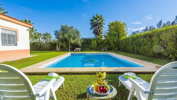 Uma piscina com água azul cristalina, rodeada por vegetação exuberante e um edifício branco com telhado em azulejos.