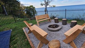 Un espace extérieur aménagé avec des bancs en bois et un foyer, entouré d’un chemin de gravier et d’une clôture en métal.