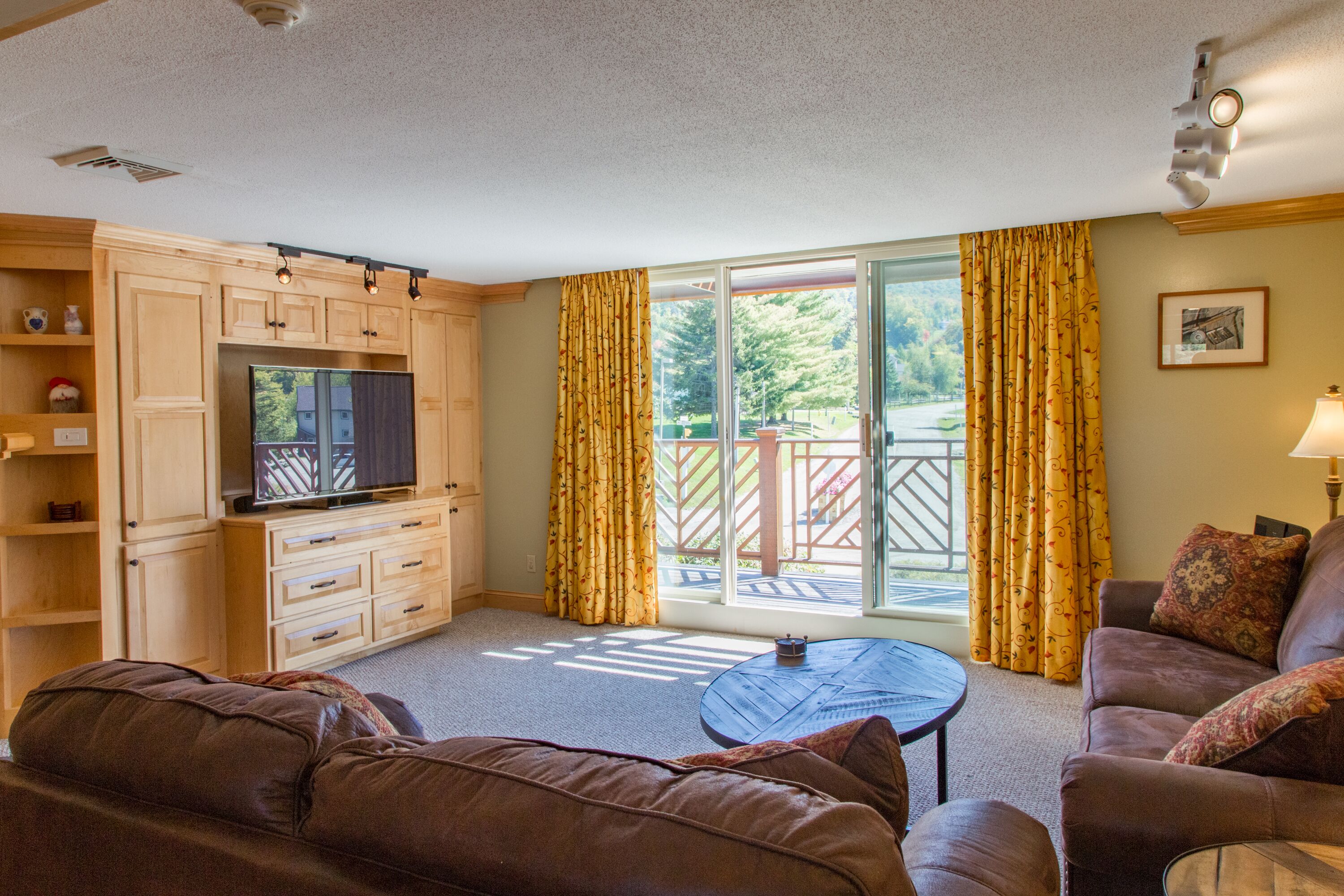 A living room with a brown leather sofa, a television, wooden cabinets, and a sliding glass door leading to an outdoor patio.