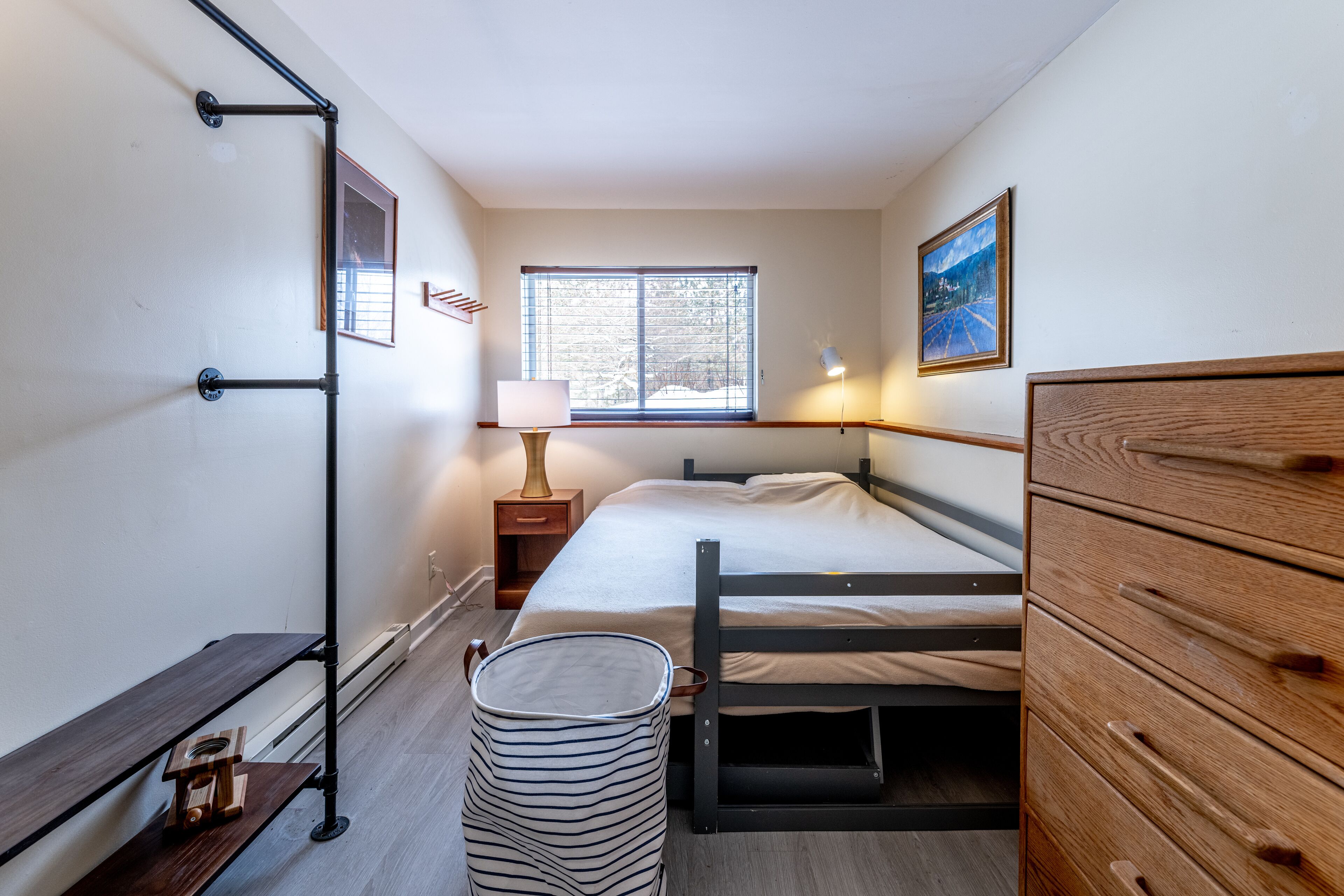 A bedroom with a bed, a nightstand, a lamp, a window, and a wooden dresser.