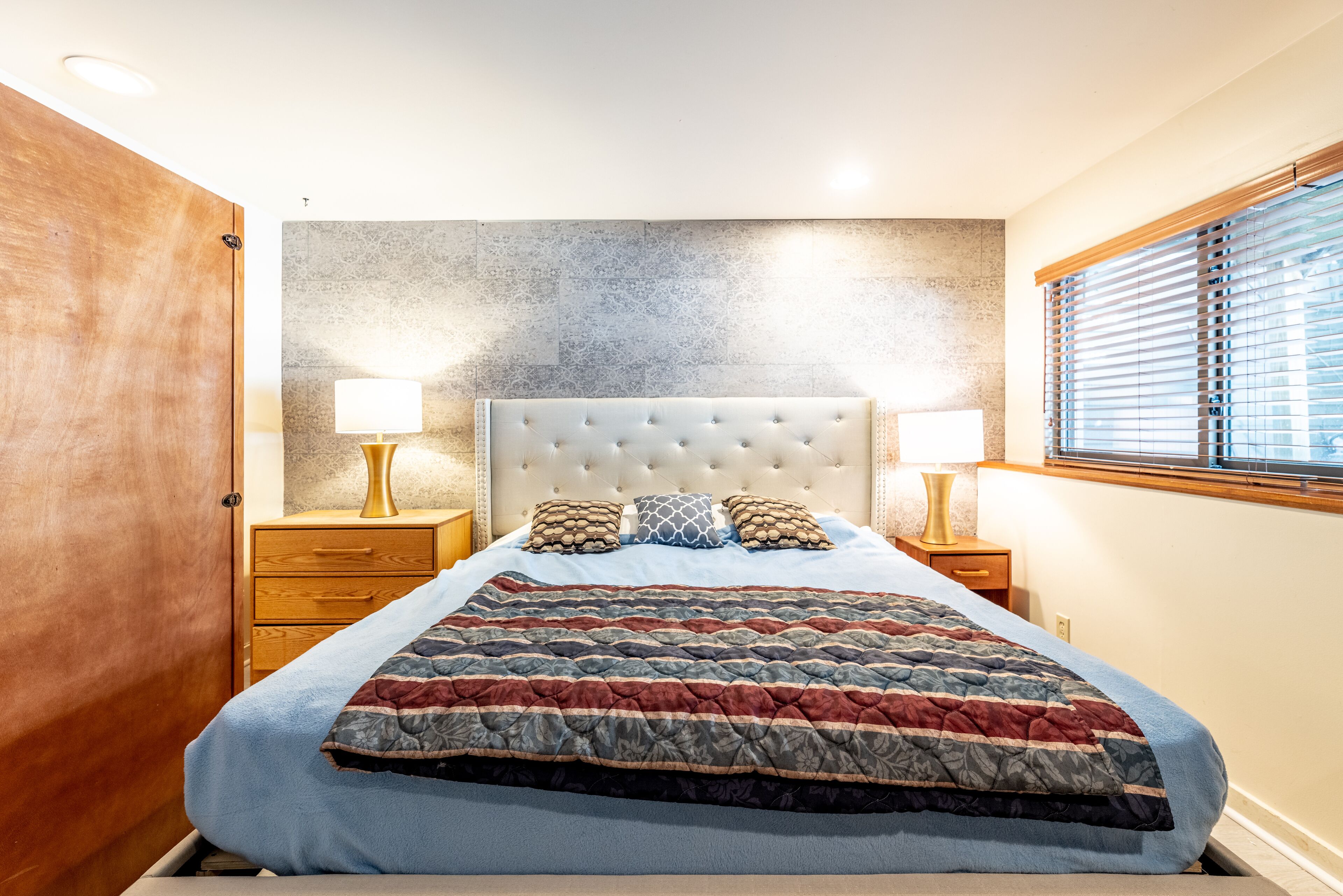 A bedroom with a tufted headboard, a quilted bedspread, bedside lamps, and a window with blinds.