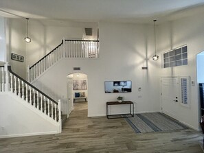 Un amplio salón con escalera, un televisor de pantalla plana en la pared y una consola con una planta.