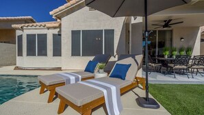Een zwembadterras met twee ligstoelen en een tafel, een parasol en uitzicht op een huis met grote ramen.