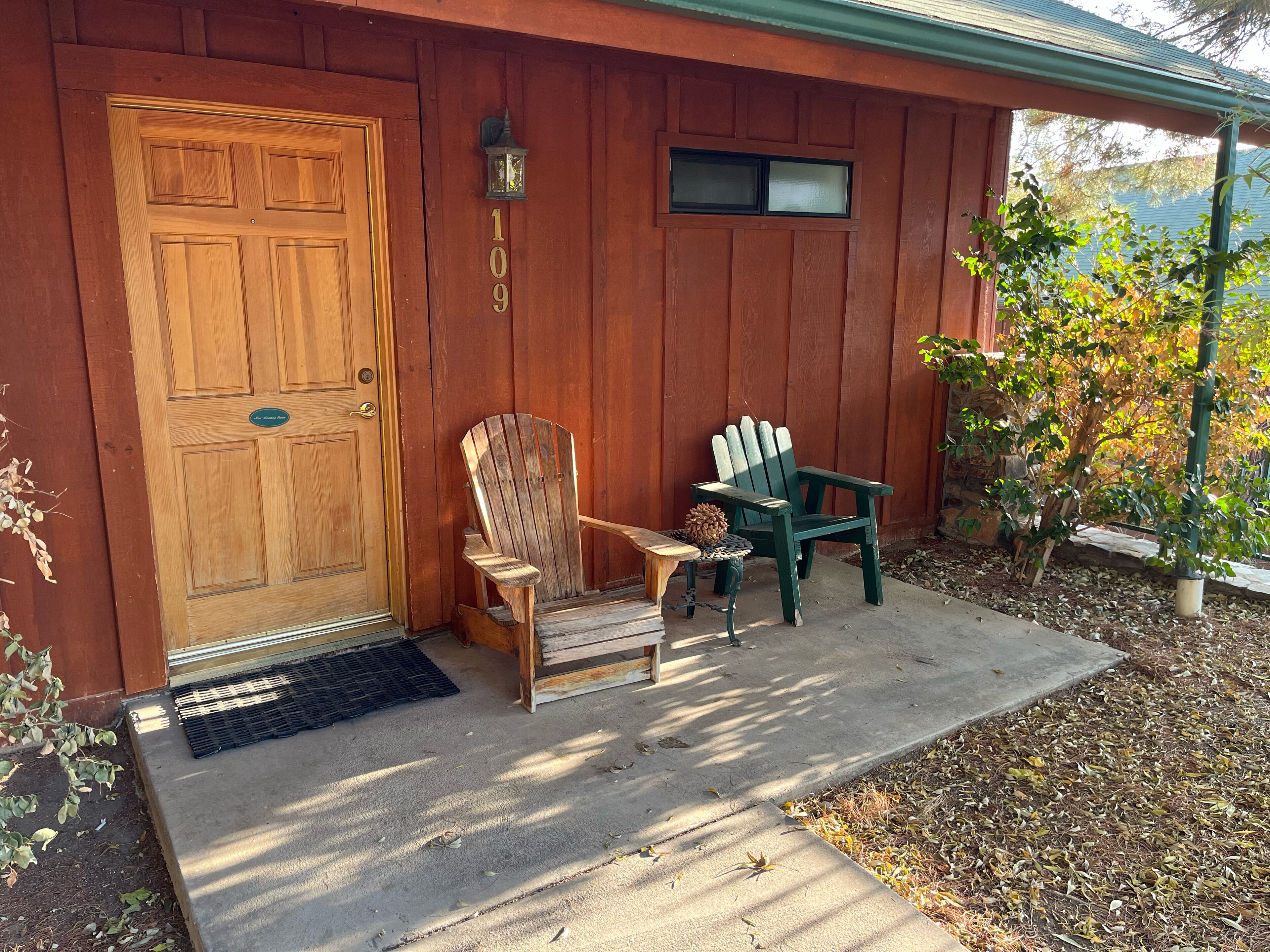 A wooden house with a double door, two Adirondack chairs, a small table, and a potted plant.