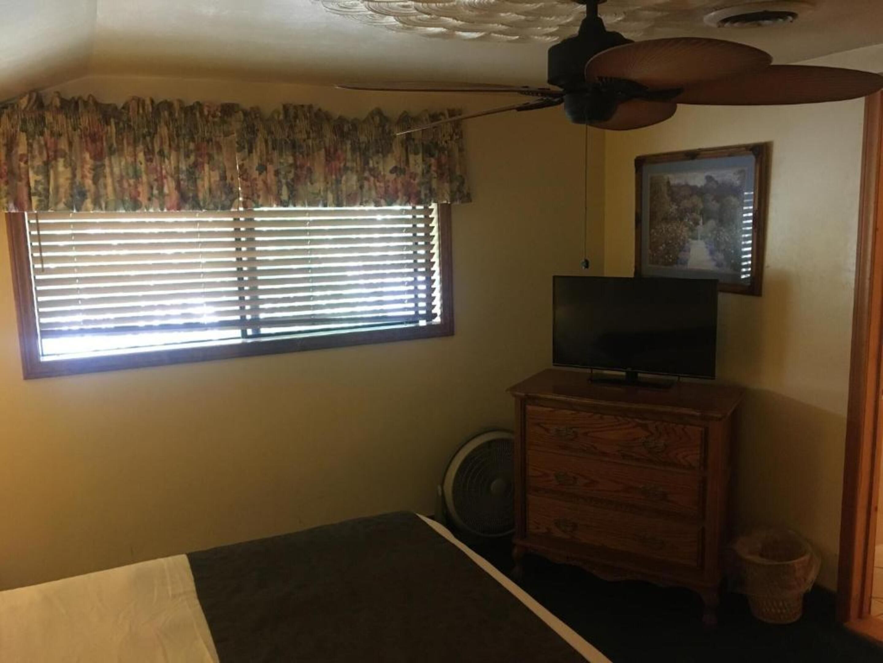 Un dormitorio con cama, cómoda, televisor, ventilador de techo y ventana con persianas.