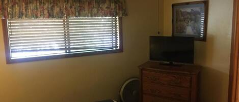 Un dormitorio con cama, cómoda, televisor, ventilador de techo y ventana con persianas.