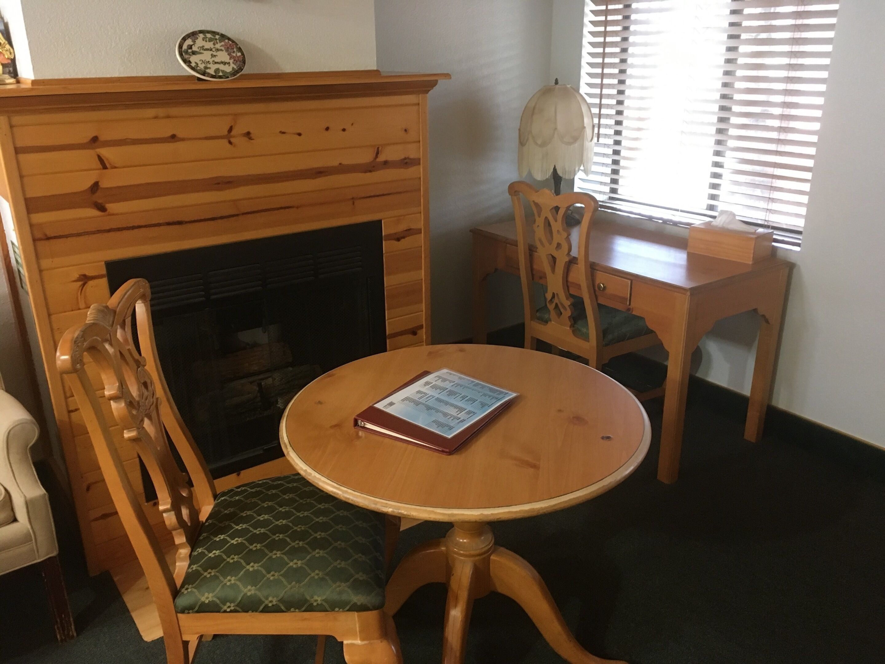 Una habitación acogedora con una chimenea de madera, una mesa redonda de madera con un menú, dos sillas de madera con cojines verdes y una ventana con persianas.