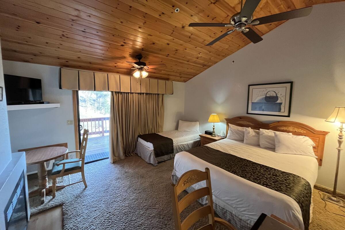 A hotel room with a wooden ceiling, two beds, a desk, a chair, a TV, a ceiling fan, and a window with curtains.