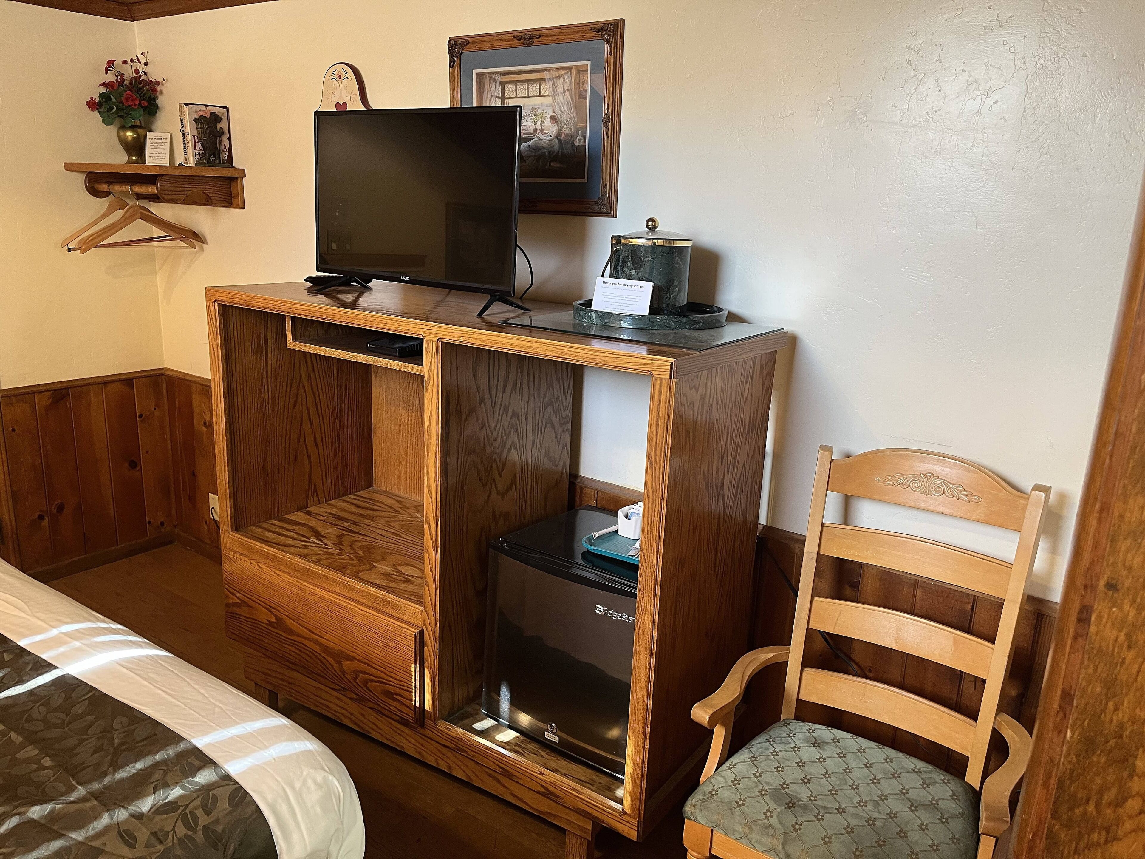 Un bureau en bois avec une télévision, une petite étagère ornée d’un vase et d’un tableau encadré, et une chaise avec un coussin.