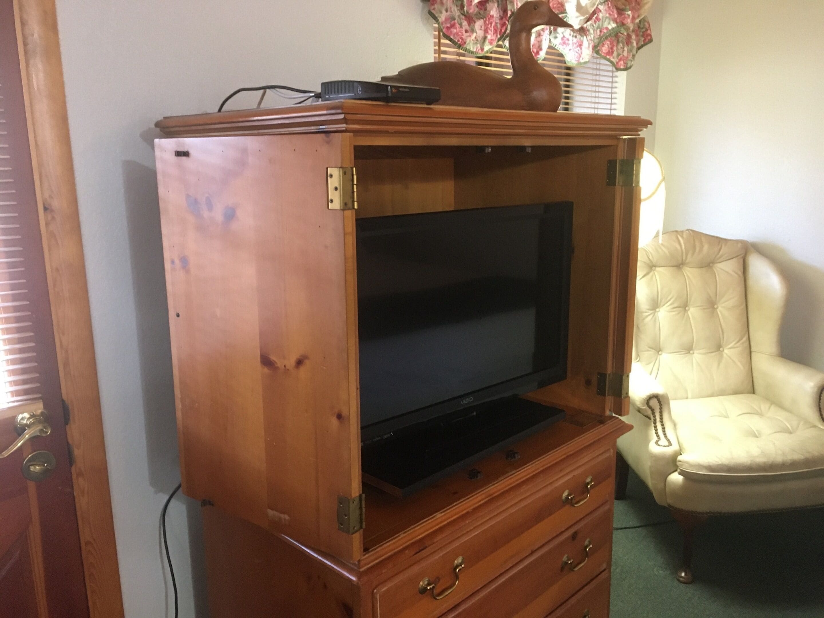 Een houten tv-meubel met een flatscreen televisie, een decoratief eendje en een gestofte fauteuil.