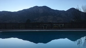 A swimming pool with a wooden deck and chairs, surrounded by mountains.