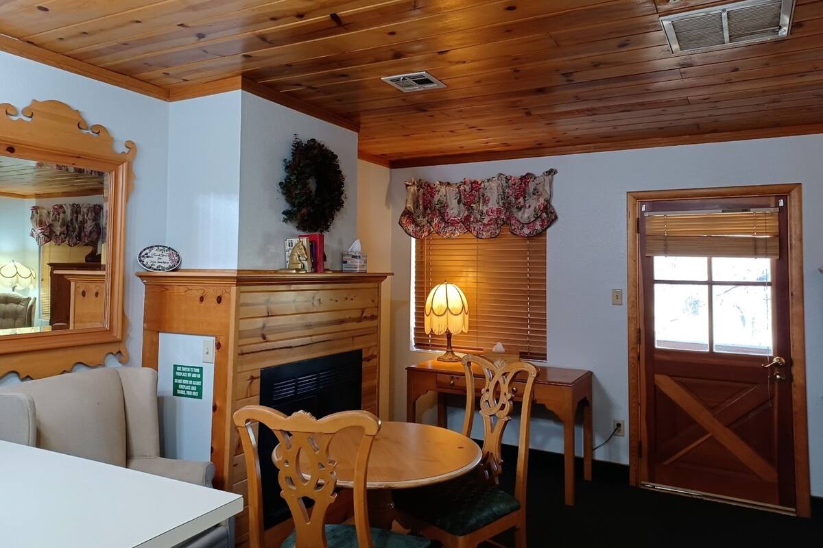 A room with wooden ceiling and walls, a fireplace with a mantel, a dining table with chairs, a door, and a window with blinds.