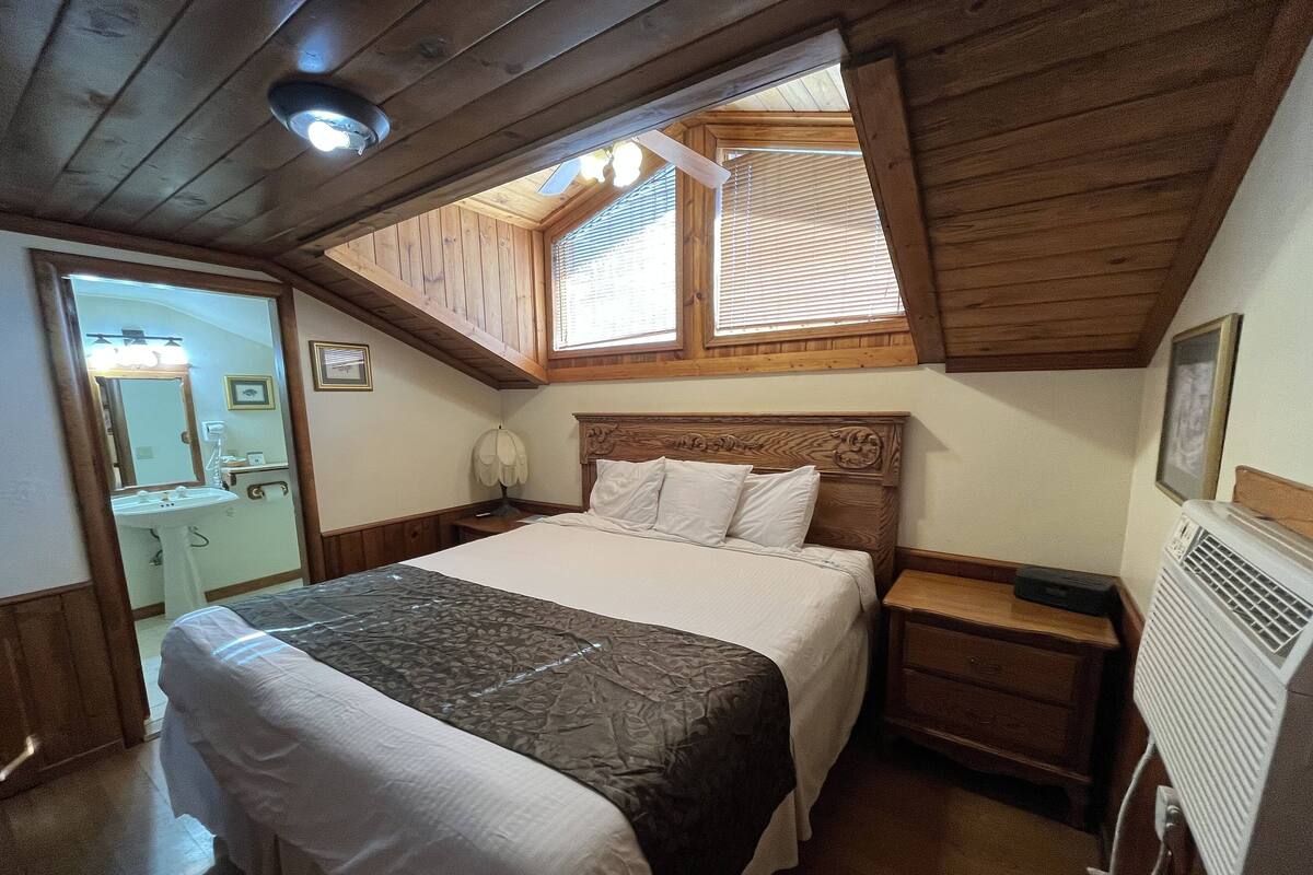 A bedroom with a wooden ceiling, a bed with white and brown bedding, a nightstand, a bedside table, a mirror, and a radiator.