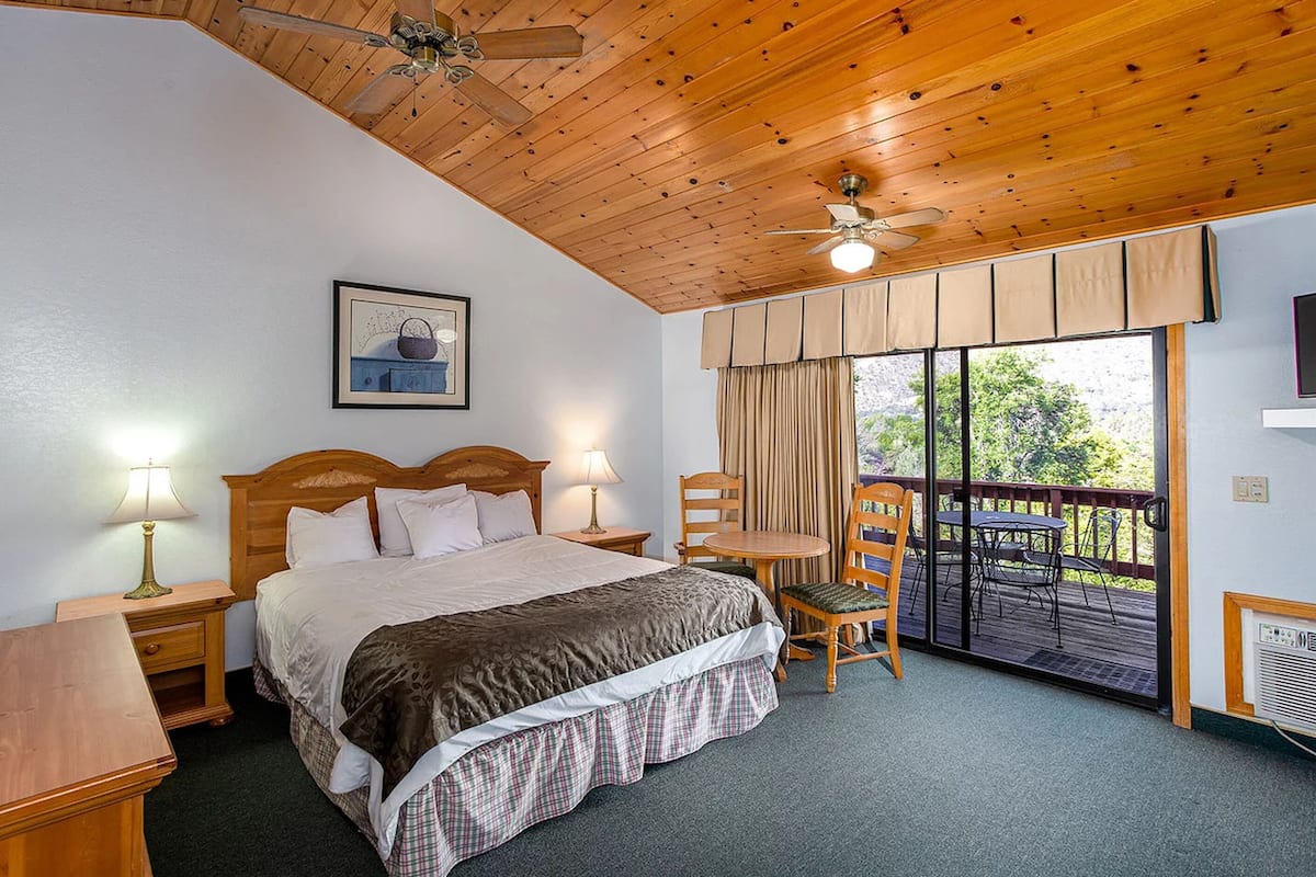 A bedroom with a wooden ceiling, a bed with a headboard, two bedside tables with lamps, a window with curtains, and a balcony with a table and chairs.
