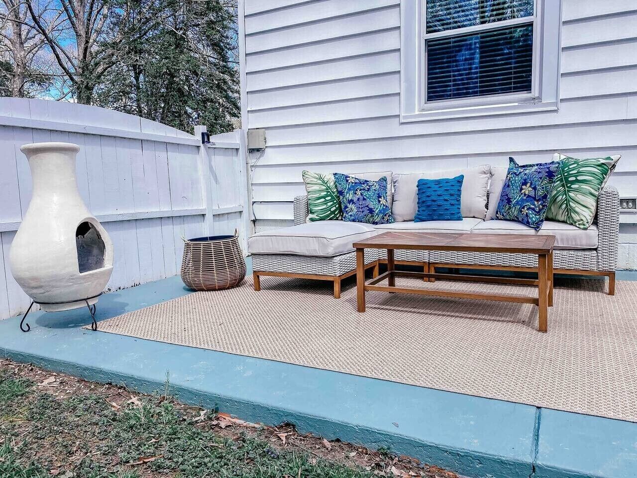 Ein Patio mit Korbusofa, Holztisch, einer großen Vase und einem Korbkorb.