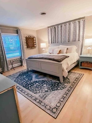 A bedroom with a bed, bedside tables, a window with curtains, a patterned rug, and a ceiling vent.