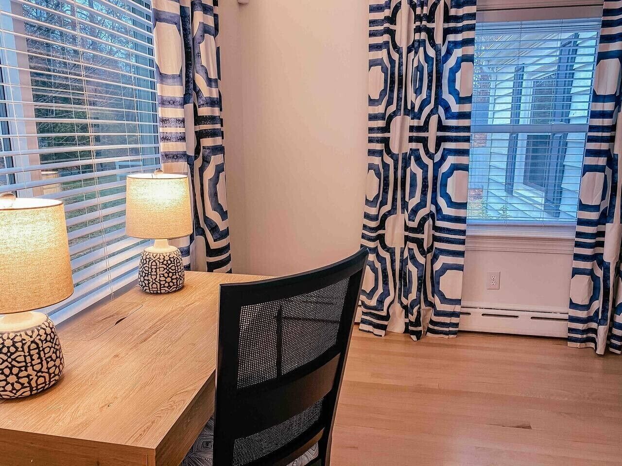 A dining area with a wooden table, two patterned lamps, a black mesh chair, and patterned curtains.