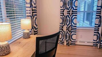 A dining area with a wooden table, two patterned lamps, a black mesh chair, and patterned curtains.