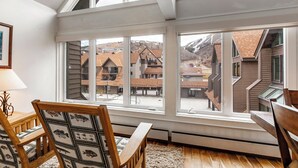 Ein Zimmer mit großen Fenstern, Holzmöbeln und Blick auf eine Stadt mit Gebäuden und Bergen.