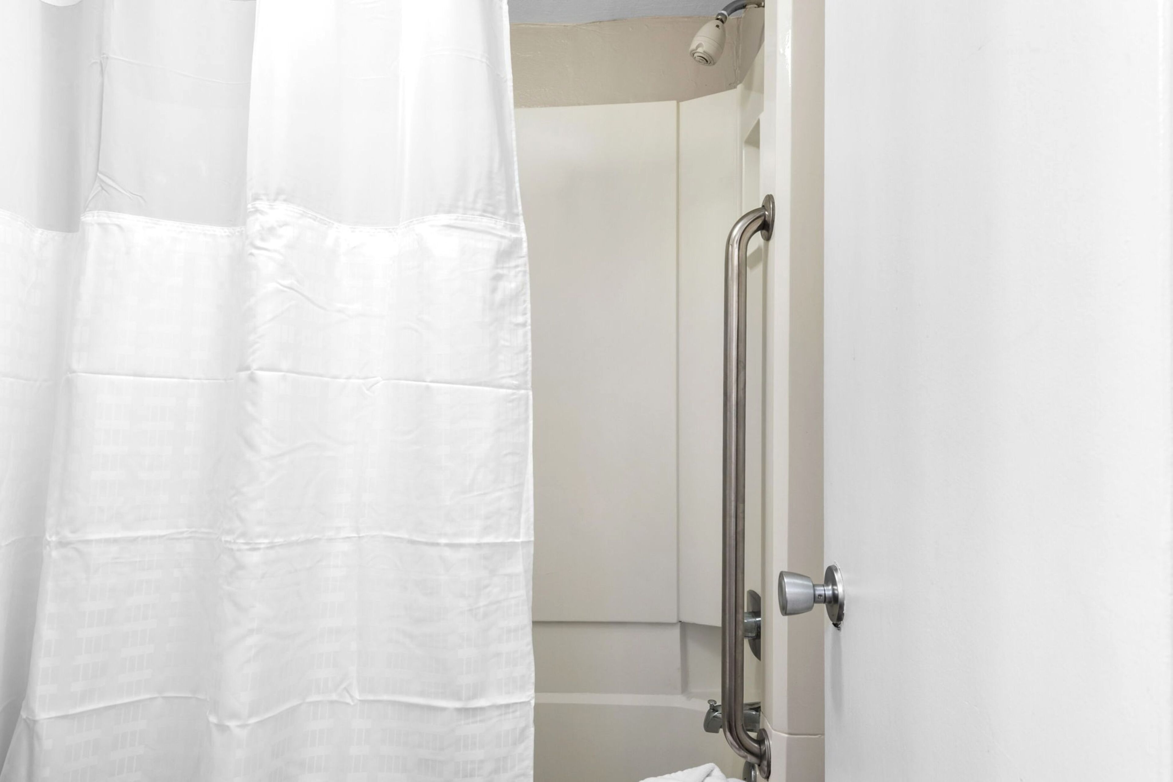 Un espace douche avec un rideau blanc, une barre d’appui et une porte dotée d’une poignée métallique.
