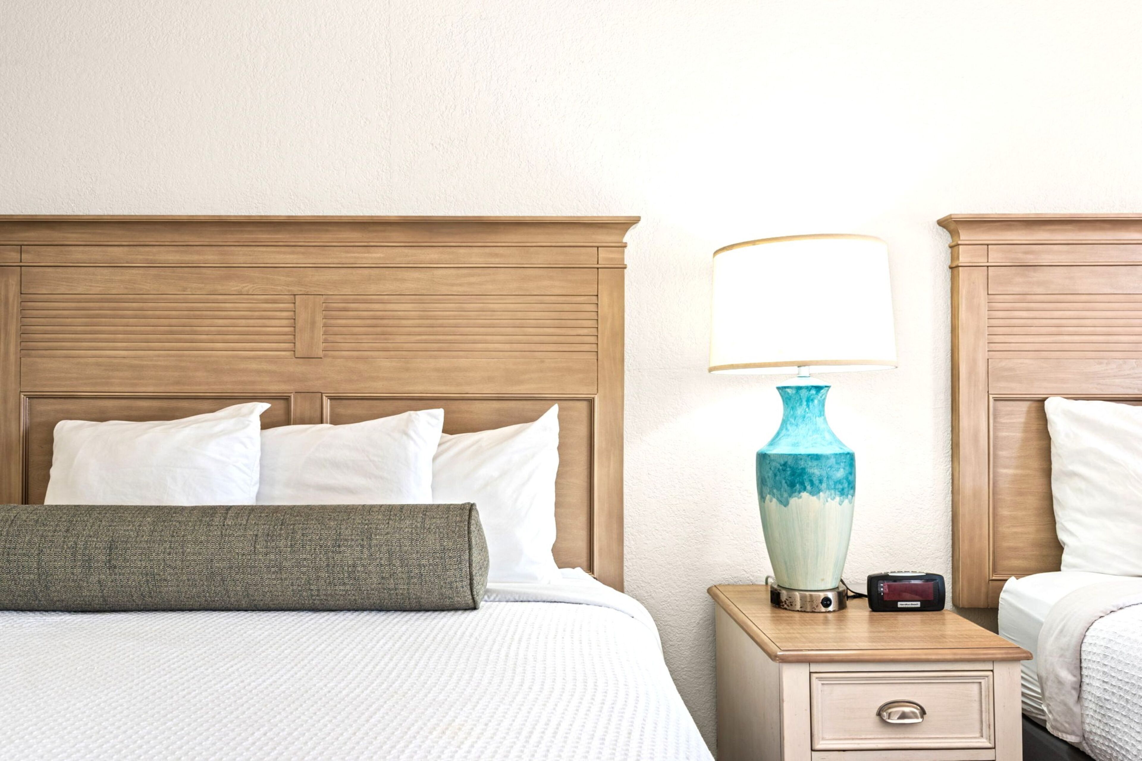 Een hotelkamer met een houten hoofdeinde, een bed met witte beddengoed, een nachtkastje met een lamp en een klein lade.