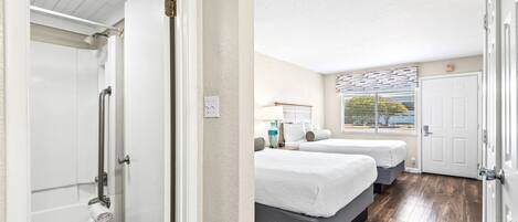 A hotel room with a shower, two beds, a window with blinds, and a door.