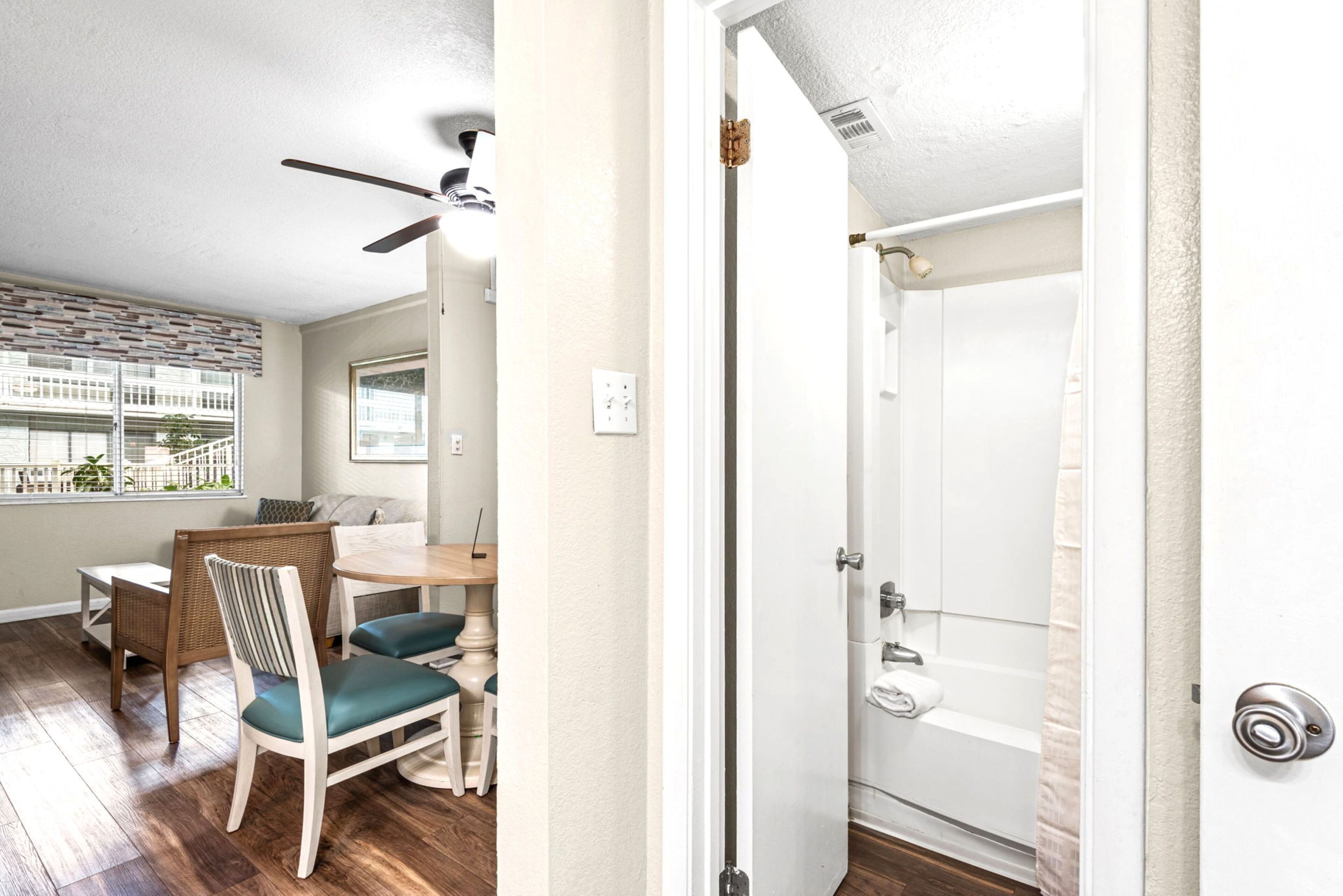 A room with a dining area, a ceiling fan, a door leading to a bathroom, and a view of a building outside.