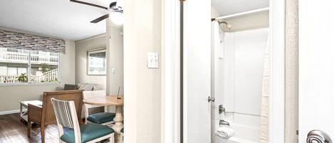 A room with a dining area, a ceiling fan, a door leading to a bathroom, and a view of a building outside.