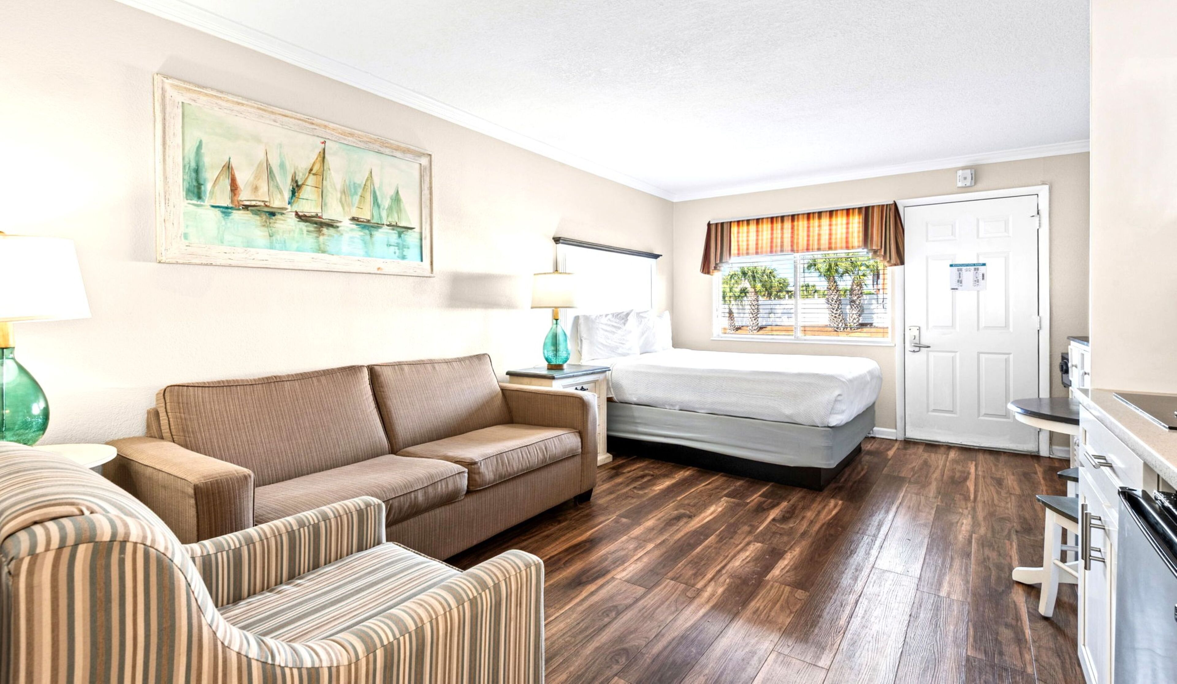 Een hotelkamer met een bed, een slaapbank, een gestreept fauteuil, een schilderij, een raam met gordijnen en een deur.