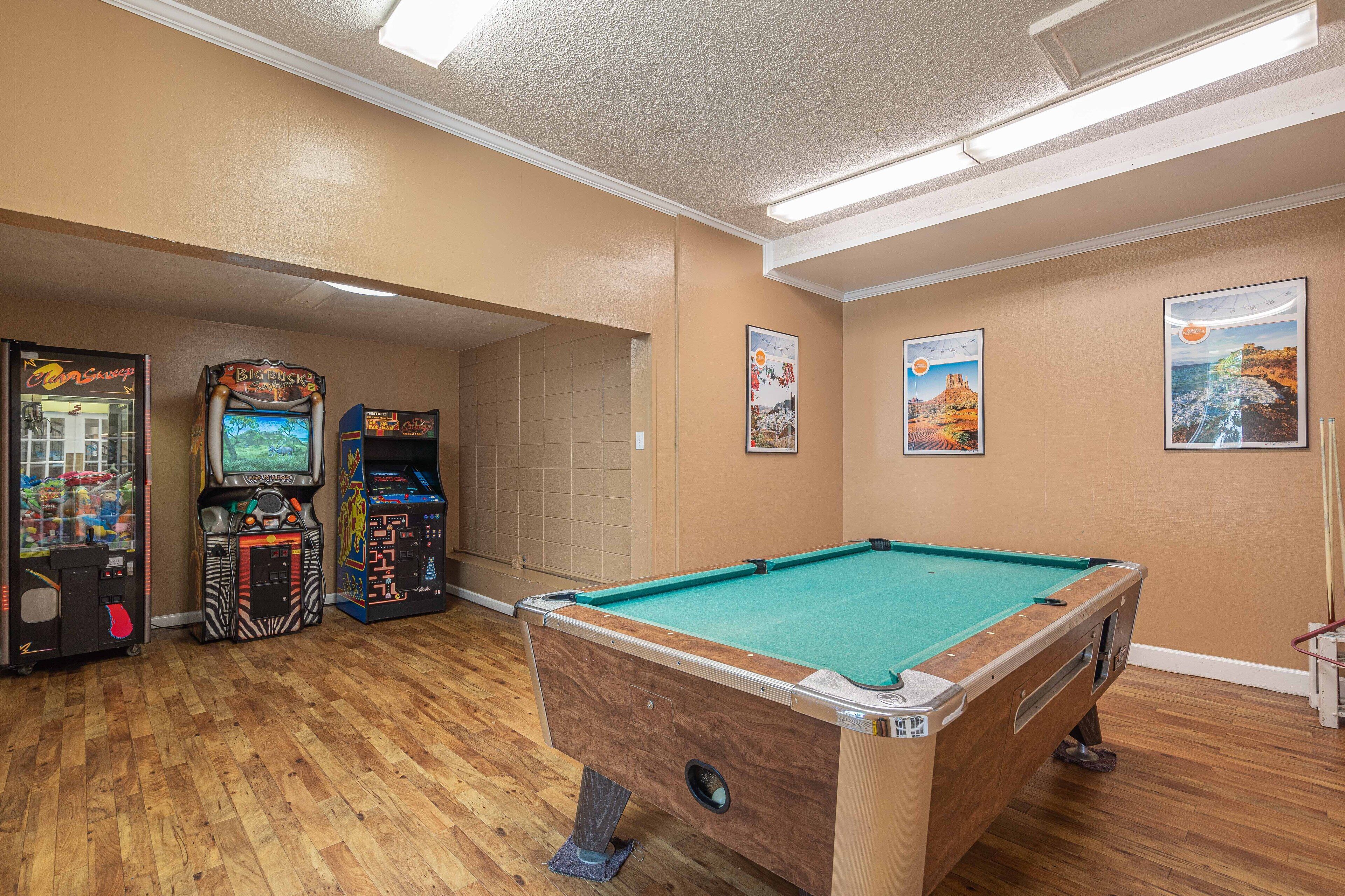 A pool table in a room with wooden flooring, arcade games, and framed pictures on the walls.