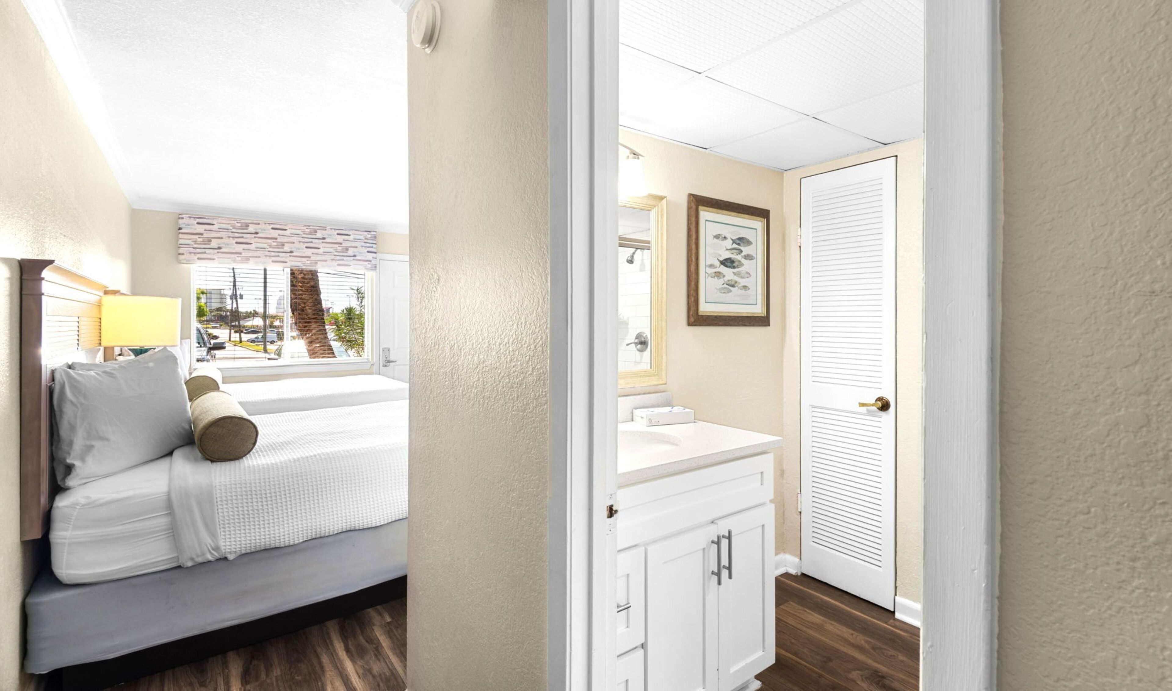 A hotel room with a bed, a window with a view of the street, a bathroom with a sink and mirror, and a door.