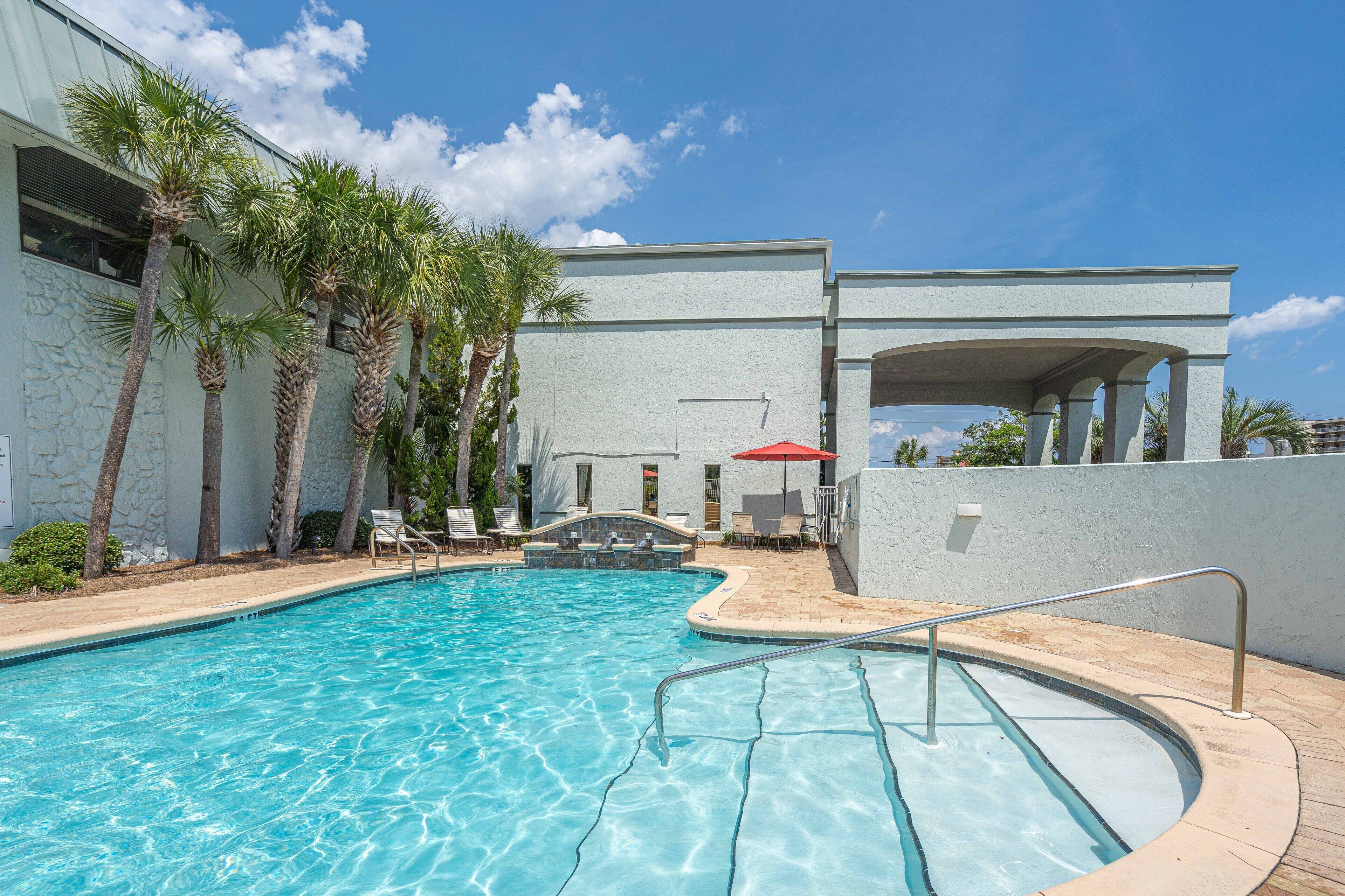 A swimming pool with clear blue water, surrounded by palm trees and a covered seating area with umbrellas.