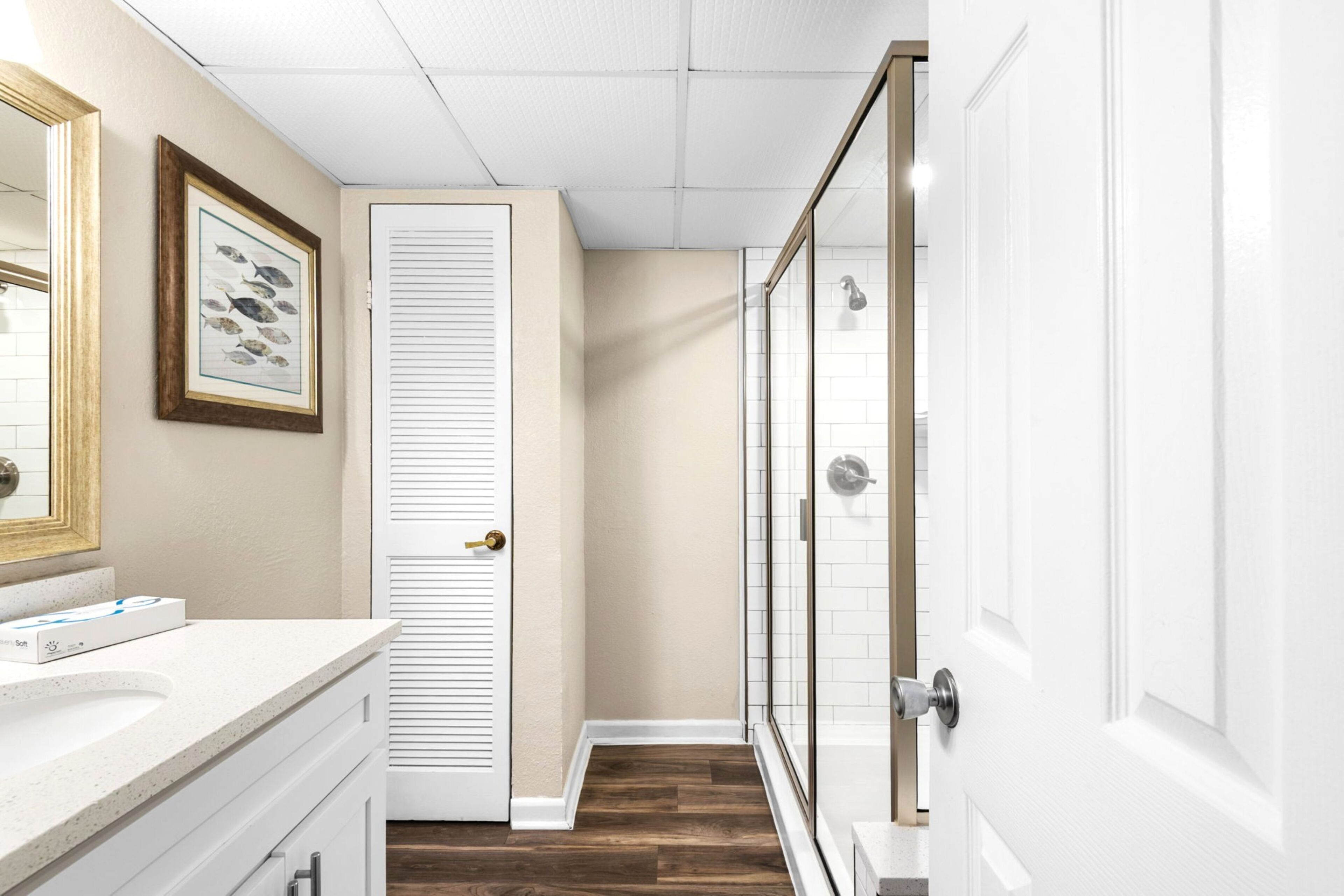 A bathroom with a white vanity, a framed fish picture, a door with a louvered design, and a glass shower enclosure.