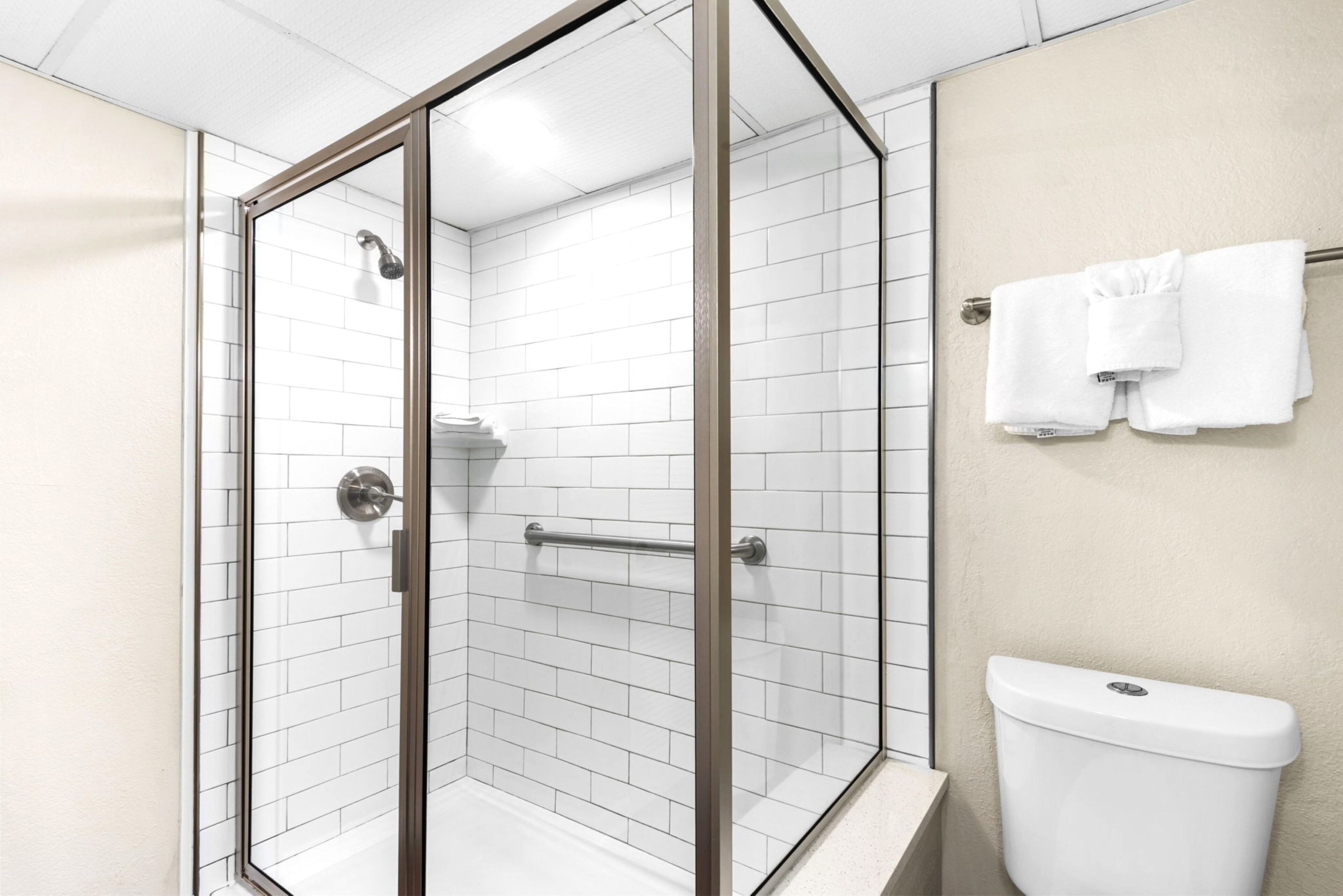 Une douche avec une porte en verre, un mur recouvert de carreaux blancs et des toilettes avec un porte-serviettes au-dessus.