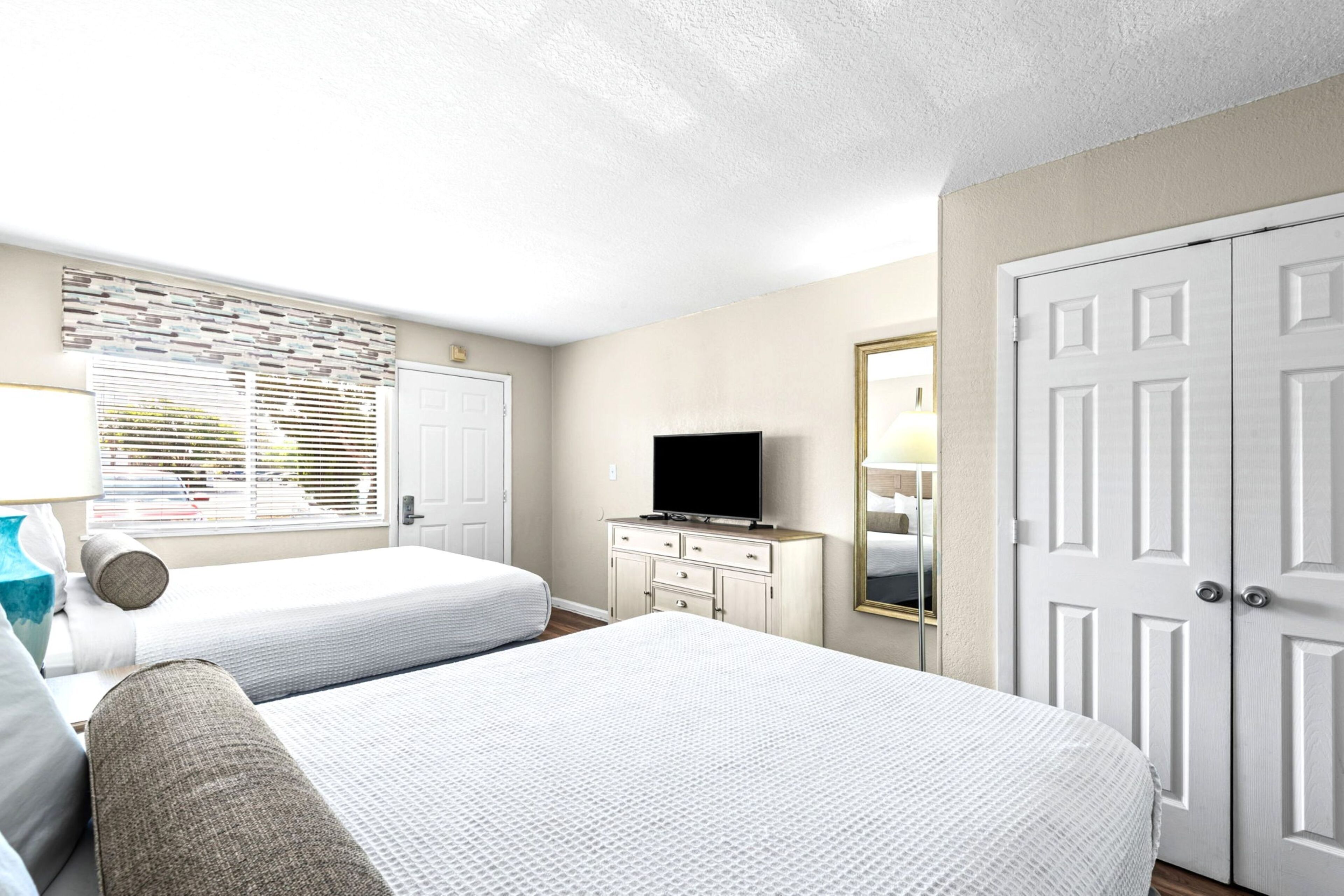 A bedroom with two beds, a dresser, a television, a window with blinds, and a door.