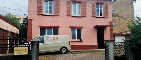 Una casa de dos pisos con fachada roja, una camioneta color beige estacionada adelante y un camino de entrada con portón.