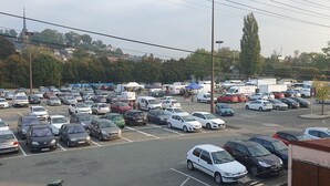 Un estacionamiento con varios autos y algunas camionetas, rodeado de árboles y edificios.