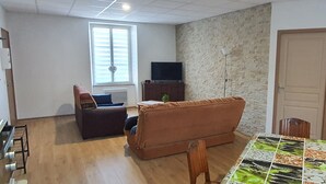 Una sala de estar con un sofá marrón, una mesita con una planta, un televisor sobre un mueble de madera y una puerta a la derecha.