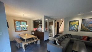 Una sala de estar moderna con un sofá gris, una mesa de comedor de madera con bancos, y una zona de cocina con electrodomésticos de acero inoxidable.