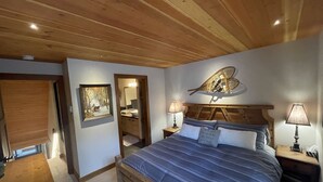 Un dormitorio con techo de madera, una cama con ropa de cama azul, una mesita de noche con lámpara, un espejo montado en la pared y un cuadro que muestra un paisaje nevado.