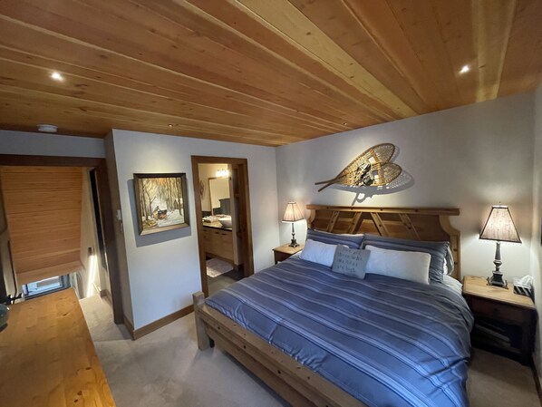 A bedroom with a wooden ceiling, a bed with blue bedding, a nightstand with a lamp, a wall-mounted mirror, and a painting of a snowy scene.