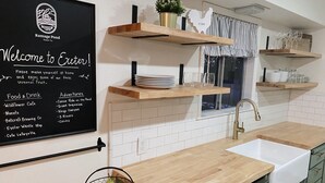 A kitchen with a blackboard listing local food and drink options, wooden shelves with glassware, and a fruit basket on the counter. - Country house with Pond and EV charger (Exeter)