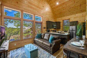 A cozy cabin interior with a wooden ceiling, large windows, a brown leather sofa, a dining table with chairs, and a kitchen area. - Cute Cabin Near PF/Island Hottub GameRoom 2Bed Sleeps 5 Air Hockey Private (Pigeon Forge)