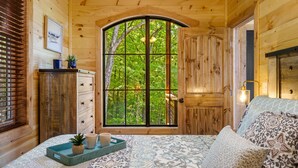 A wooden cabin bedroom with a large arched window, a bed with patterned bedding, a bedside table with a lamp, and a small plant on the windowsill. - Adorable Cabin Near PF/Island Hottub GameRoom 2Bed Sleeps 5 Air Hockey Private (Pigeon Forge)
