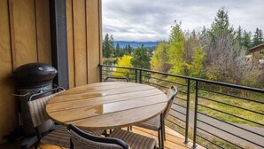 Eine Holzterrasse mit rundem Tisch und Stühlen, einem Grill und Blick auf Bäume und Berge.