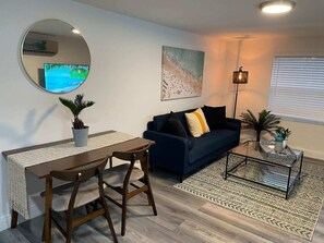 A living room with a round mirror, a blue sofa, a coffee table, and a dining area with a table and chairs. - Newly refurbished apartment with private garden (Fort Lauderdale)