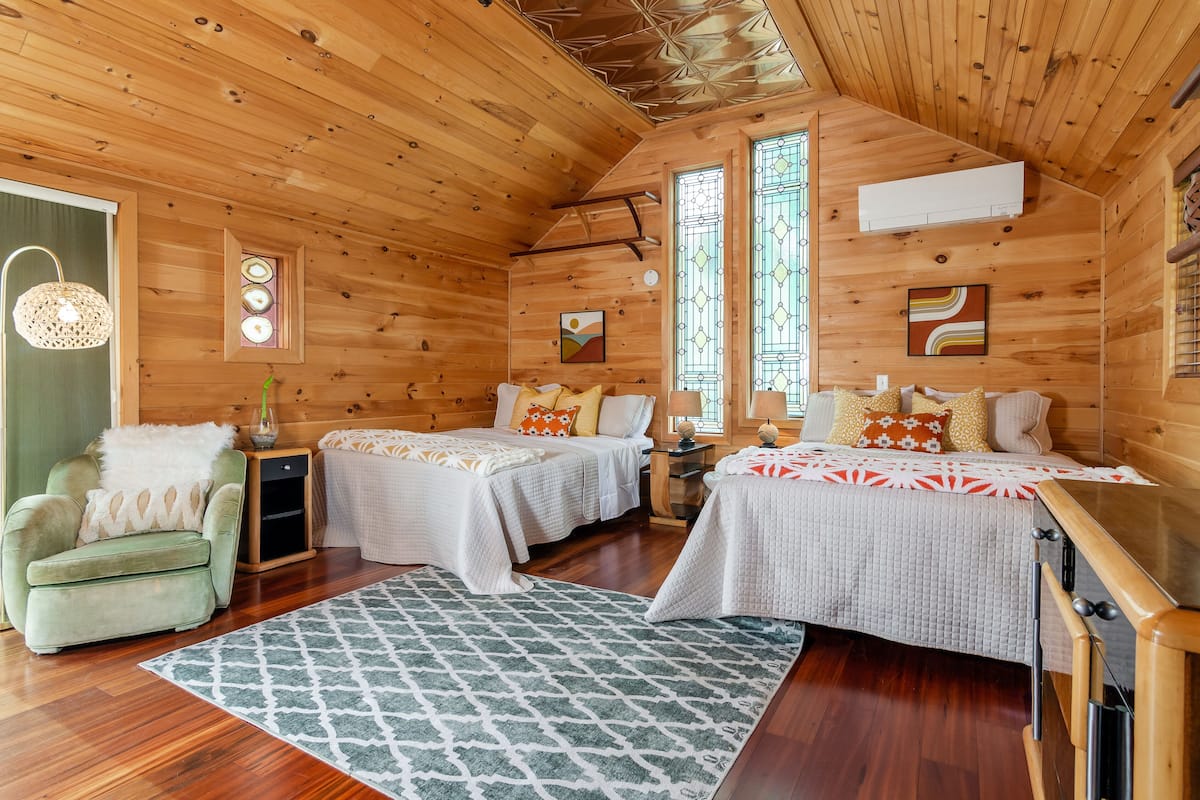 A wooden cabin with two beds, a green armchair, and a patterned rug.